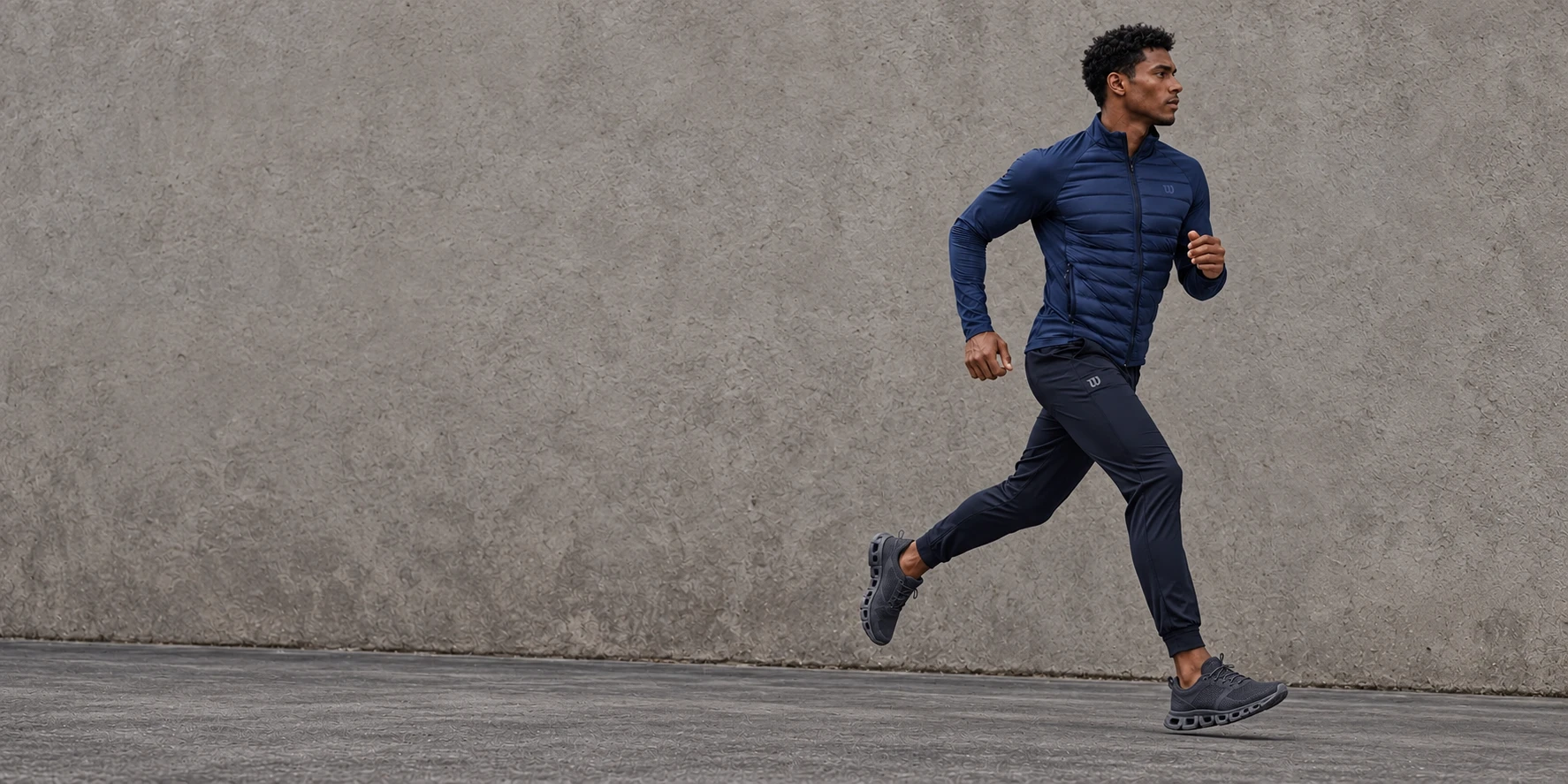 Athlete running in a navy Wilson jacket, dark pants and unbranded charcoal shoes against a stone-gray wall
